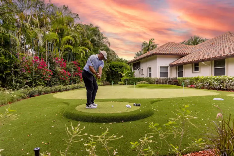 A golfer practicing short game shots on a custom backyard synthetic putting green using a dedicated chipping mat at sunset outside a luxury Belleair, Florida home.