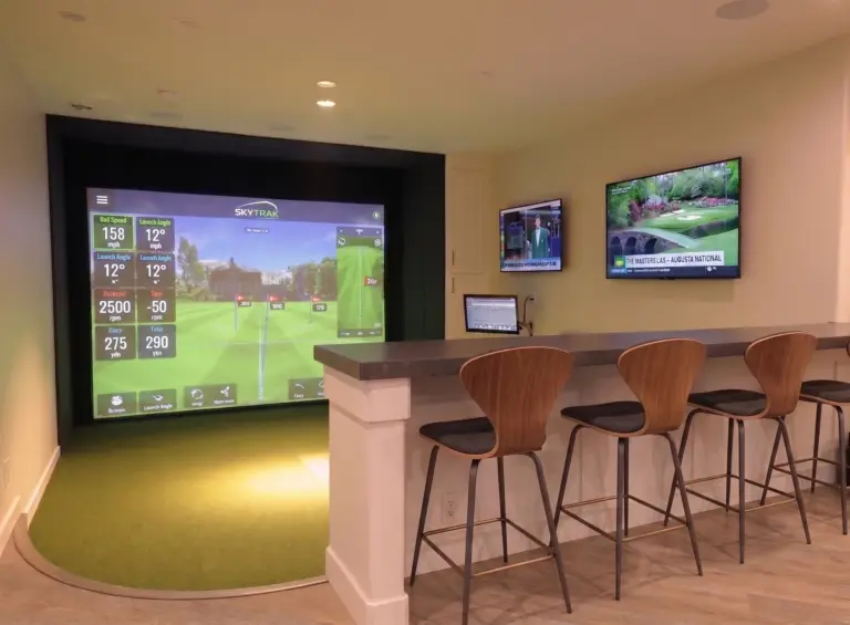 A custom-engineered luxury residential indoor golf simulator sanctuary installation featuring a seamless, flush-mounted synthetic turf flooring that transitions perfectly from the putting mat to the large impact screen, shown in a private home setting.