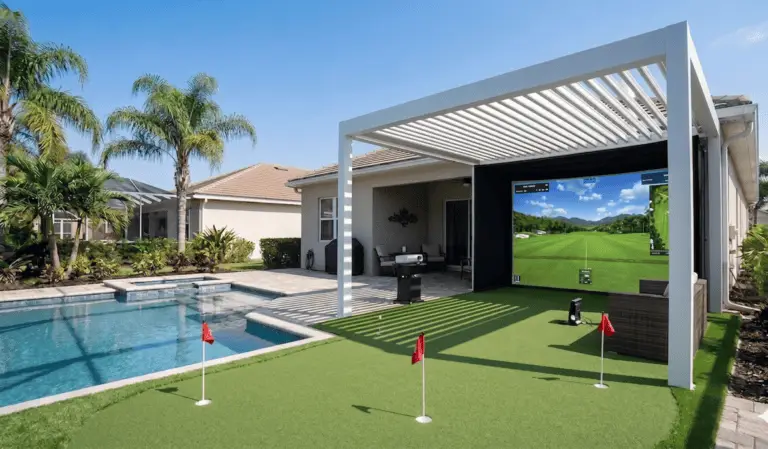 A bright outdoor patio featuring a white louvered pergola covering a large golf simulator screen. In front of the screen is a custom artificial putting green with three red