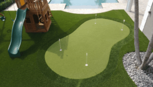 Custom Backyard Putting Green and Playground in Madeira Beach, Florida - Sims & Greens - Sims & Greens Elevated view of a custom artificial turf putting green with four holes and white flags, located next to a wooden playground structure with a green slide, with a modern swimming pool in the background.