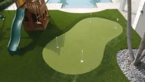 Custom Backyard Putting Green and Playground in Madeira Beach, Florida - Sims & Greens - Sims & Greens Elevated view of a custom artificial turf putting green with four holes and white flags, located next to a wooden playground structure with a green slide, with a modern swimming pool in the background.