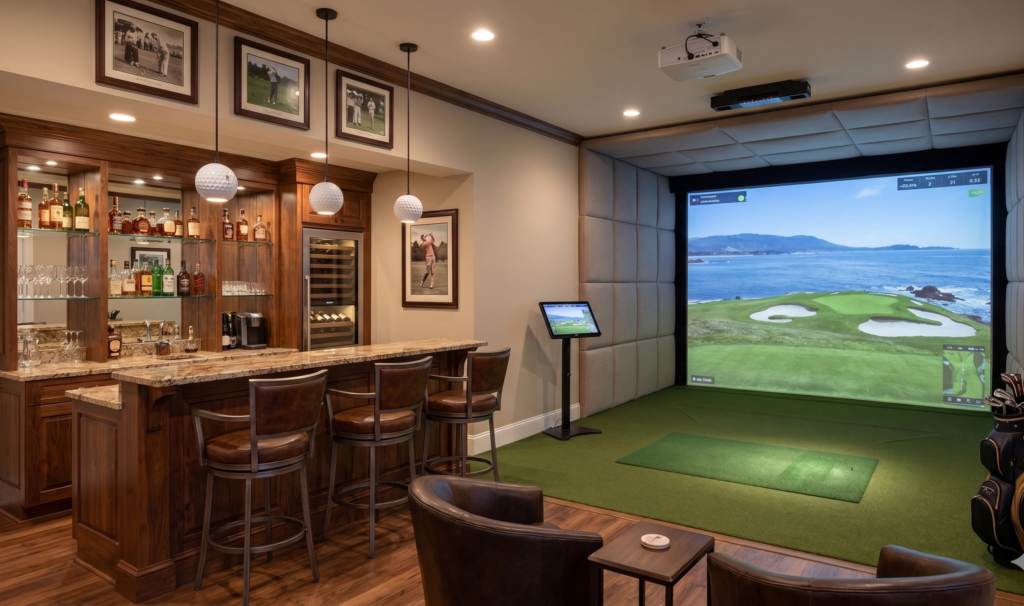 A luxurious home entertainment room featuring a custom dark wood wet bar with three leather barstools on the left, and a professional indoor golf simulator bay on the right. The simulator has a large screen projecting a coastal golf course, surrounded by padded walls and green turf. Two leather lounge chairs sit in the foreground on a hardwood floor.