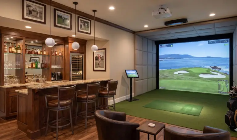 A luxurious home entertainment room featuring a custom dark wood wet bar with three leather barstools on the left, and a professional indoor golf simulator bay on the right. The simulator has a large screen projecting a coastal golf course, surrounded by padded walls and green turf. Two leather lounge chairs sit in the foreground on a hardwood floor.