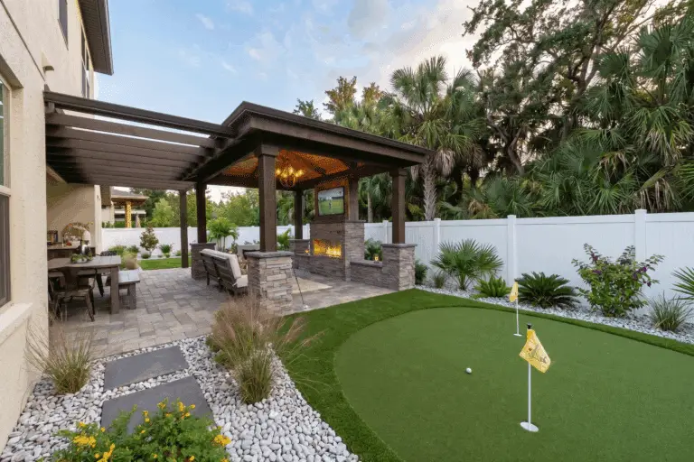 A modern backyard in Tampa, Florida, featuring a light tan stucco house, a professional synthetic putting green with two flags, and a large dark wood pavilion with a stone fireplace and TV. The garden beds are filled with white river stones and Florida native plants like Muhly grass and Saw Palmettos, all enclosed by a white PVC privacy fence.