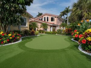 Clearwater-artif - Sims & Greens Residential backyard with a curved artificial putting green, tropical plants, and a pink house in the background on a sunny day by the sea of greenery.