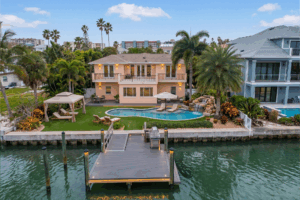Two-story peach villa by the water with a balcony, pool, and palm trees in a tropical backyard setting near other houses on the canal.