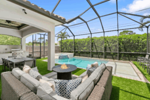 Outdoor screened patio with wicker sectional seating, fire pit table, dining area, and a rectangular pool bordered by green hedges under a metal frame enclosure.