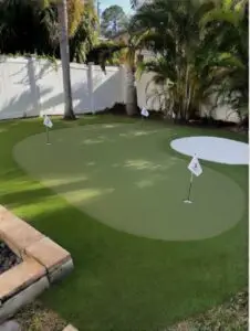 Backyard Putting Green with White Turf Bunker – Seminole, FL - Sims & Greens Professional backyard putting green installation in Seminole, Florida with curved light green artificial turf, a distinctive white turf bunker, and multiple white practice flags. Palm trees and white vinyl fencing visible in the sunny residential backyard setting.