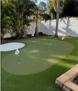 Professional backyard putting green installation in Seminole, Florida with curved light green artificial turf, a distinctive white turf bunker, and multiple white practice flags. Palm trees and white vinyl fencing visible in the sunny residential backyard setting.