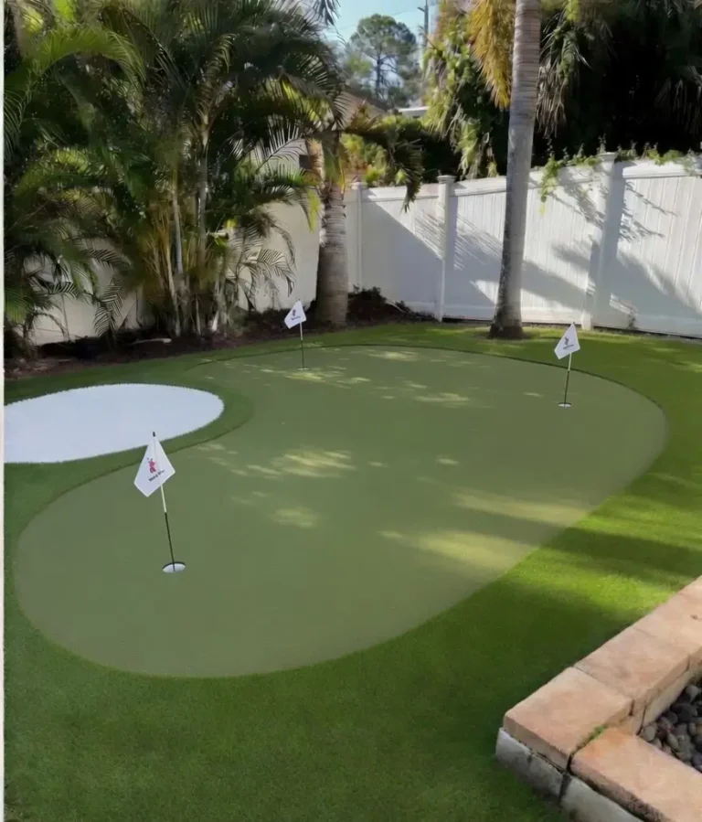 Professional backyard putting green installation in Seminole, Florida with curved light green artificial turf, a distinctive white turf bunker, and multiple white practice flags. Palm trees and white vinyl fencing visible in the sunny residential backyard setting.
