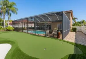 palm-harbor-backyard-putting=green-bunker - Sims & Greens Custom artificial turf backyard putting green with an integrated white sand bunker next to a screened-in pool enclosure and palm trees in Palm Harbor, Florida.